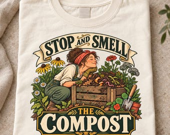 May include: A cream-colored t-shirt featuring a graphic of a person kneeling by a wooden compost bin, surrounded by flowers and a small shovel. The text reads STOP AND SMELL THE COMPOST in bold, dark lettering with a decorative banner design.