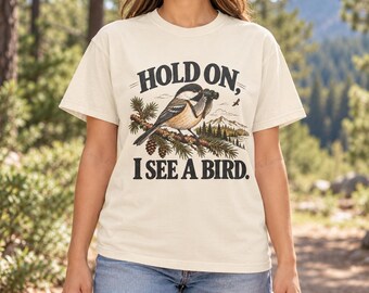 May include: A cream-colored short-sleeve t-shirt featuring a graphic of a bird perched on a pine branch holding binoculars. The text "HOLD ON, I SEE A BIRD." is printed in black above and below the illustration of the bird and mountains.