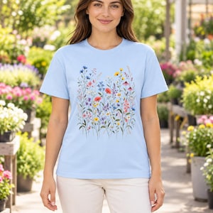 May include: A light blue short-sleeve t-shirt featuring a central graphic print of various colorful wildflowers including red poppies, yellow daisies, and blue cornflowers arranged in a vertical botanical pattern on the front.