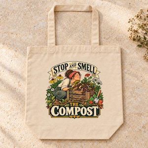 May include: A beige canvas tote bag featuring a colorful graphic of a person kneeling by a wooden compost bin. The text reads STOP AND SMELL THE COMPOST. The illustration includes various flowers, green foliage, and a small garden trowel.