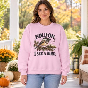 May include: A light pink crewneck sweatshirt featuring a graphic of a chickadee bird holding binoculars on a pine branch with a mountain landscape. The text reads, "HOLD ON, I SEE A BIRD." The sweatshirt has long sleeves and a ribbed neckline.