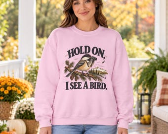 May include: A light pink crewneck sweatshirt featuring a graphic of a chickadee bird holding binoculars on a pine branch with a mountain landscape. The text reads, "HOLD ON, I SEE A BIRD." The sweatshirt has long sleeves and a ribbed neckline.