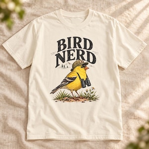 May include: An off-white short-sleeve t-shirt featuring a graphic of a yellow bird wearing a green cap and carrying black binoculars. The text BIRD NERD is printed in black above the bird, which stands in green grass with small flowers.