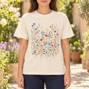May include: A cream-colored short-sleeve t-shirt featuring a central graphic print of various wildflowers in shades of red, blue, yellow, and purple, with green stems and leaves arranged in a vertical botanical pattern across the chest.