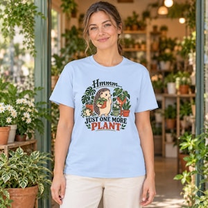 May include: A light blue short-sleeve t-shirt featuring a graphic of a hedgehog holding a potted plant surrounded by various houseplants. The text reads "Hmmm... JUST ONE MORE PLANT" in dark green and red lettering on the front of the shirt.