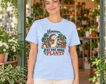 May include: A light blue short-sleeve t-shirt featuring a graphic of a hedgehog holding a potted plant surrounded by various houseplants. The text reads "Hmmm... JUST ONE MORE PLANT" in dark green and red lettering on the front of the shirt.