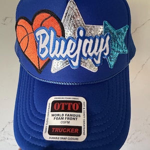 Puede incluir: Gorra de camionero azul con la palabra "Bluejays" en escritura blanca. La gorra presenta un corazón de baloncesto, una estrella de lentejuelas y una estrella azul de lentejuelas. La gorra tiene una parte delantera de espuma y un cierre a presión.
