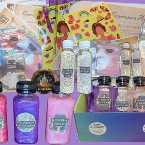 May include: A collection of colorful bottles, jars, and packets, with labels in Spanish. The bottles contain pink and purple liquids, and white spherical objects. The box includes stickers, cards, and a decorative round emblem.