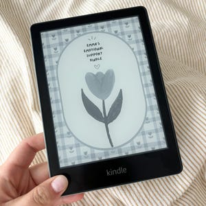 May include: A black Kindle e-reader displaying a grayscale image with the text "Emma's Emotional Support Kindle" and a floral design. The device is held against a striped beige background.