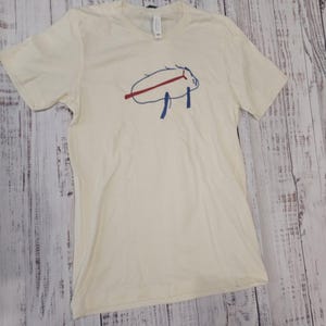 Può includere: T-shirt color crema con un design grafico blu e rosso. La grafica sembra essere un animale o un oggetto stilizzato. La maglietta è realizzata in un materiale morbido e ha maniche corte.