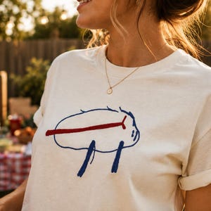May include: A cream-colored short-sleeve t-shirt featuring a central graphic design. The graphic consists of a blue hand-drawn style outline of a buffalo shape with a horizontal red line crossing through the middle and two blue vertical lines below.