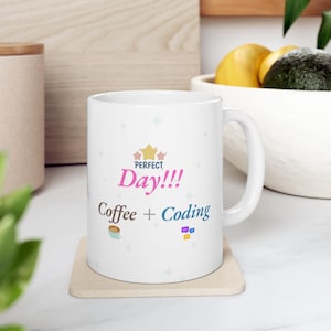 Puede incluir: Taza de cerámica blanca con el texto "Perfect Day!!! Coffee + Coding" en rosa, marrón y azul. La taza tiene un asa y está sobre un posavasos beige. La taza está decorada con formas de estrella.