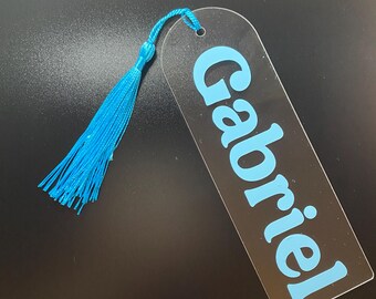 Puede incluir: Un marcapáginas de acrílico transparente con un borla azul. El marcapáginas tiene el nombre "Gabriel" impreso en azul.
