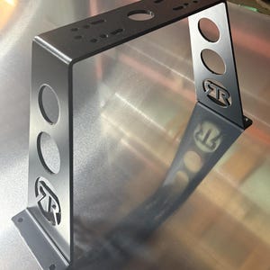 Puede incluir: Un soporte met&aacute;lico negro con una parte superior rectangular y dos soportes verticales. Los soportes tienen tres recortes circulares y un logotipo estilizado. La parte superior tiene m&uacute;ltiples agujeros para el montaje. El soporte est&aacute; dise&ntilde;ado para soporte estructural.