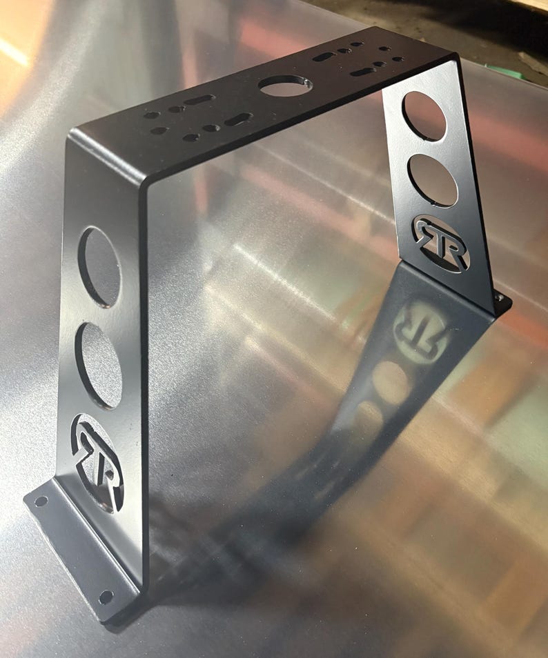 Puede incluir: Un soporte met&aacute;lico negro con una parte superior rectangular y dos soportes verticales. Los soportes tienen tres recortes circulares y un logotipo estilizado. La parte superior tiene m&uacute;ltiples agujeros para el montaje. El soporte est&aacute; dise&ntilde;ado para soporte estructural.