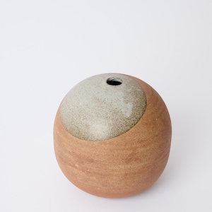 Puede incluir: Un jarrón de cerámica esférico con un diseño de dos tonos. La parte inferior es de un marrón cálido, mientras que la sección superior es de un gris claro moteado. El jarrón tiene una sola abertura circular en la parte superior. Adecuado para la decoración del hogar.