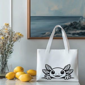 K&ouml;nnte beinhalten: Eine wei&szlig;e Tragetasche mit einer schwarzen Strichgrafik eines Axolotls, der &uuml;ber den unteren Rand schaut. Das Design zeigt das Tier mit gro&szlig;en Augen und gefiederten &auml;u&szlig;eren Kiemen, deutlich auf die Vorderseite der glatten wei&szlig;en Stofftasche gedruckt.