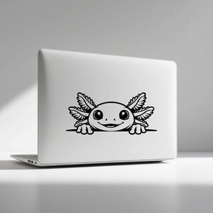 K&ouml;nnte beinhalten: Ein silberner Laptop mit einem schwarzen Vinyl-Aufkleber eines Cartoon-Axolotls. Das Axolotl ist mit gro&szlig;en Augen, einem l&auml;chelnden Mund und gefiederten &auml;u&szlig;eren Kiemen dargestellt und scheint &uuml;ber eine horizontale Linie auf dem Deckel zu schauen.