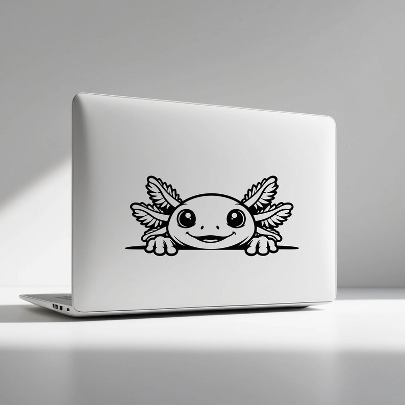 K&ouml;nnte beinhalten: Ein silberner Laptop mit einem schwarzen Vinyl-Aufkleber eines Cartoon-Axolotls. Das Axolotl ist mit gro&szlig;en Augen, einem l&auml;chelnden Mund und gefiederten &auml;u&szlig;eren Kiemen dargestellt und scheint &uuml;ber eine horizontale Linie auf dem Deckel zu schauen.