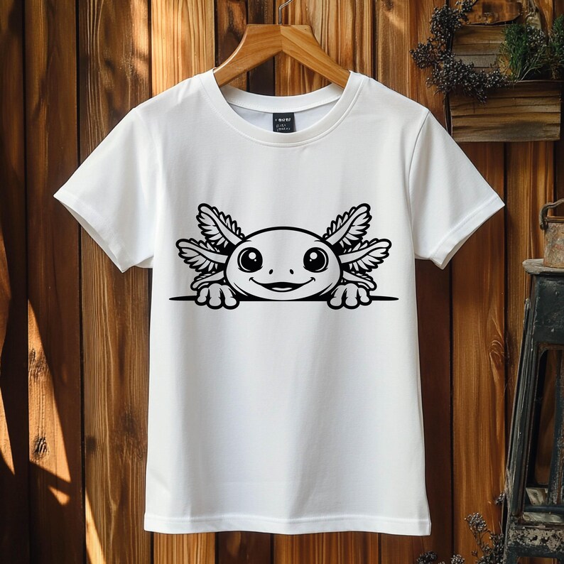 K&ouml;nnte beinhalten: Ein wei&szlig;es Kurzarm-T-Shirt mit einem schwarzen Grafikdruck eines Axolotls, das &uuml;ber einer horizontalen Linie hervorlugt. Das Axolotl hat gro&szlig;e runde Augen, ein breites L&auml;cheln und markante federartige Kiemen an beiden Kopfseiten.