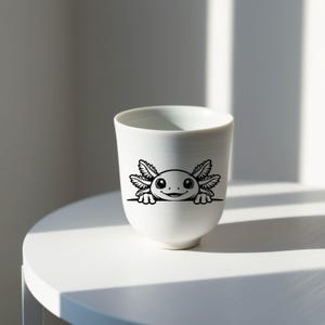 K&ouml;nnte beinhalten: Ein wei&szlig;er Keramikbecher mit einer schwarzen Strichzeichnung eines Axolotls, der &uuml;ber eine horizontale Linie schaut. Der Axolotl hat gro&szlig;e Augen, einen kleinen Mund und auff&auml;llige, federartige &auml;u&szlig;ere Kiemen an beiden Seiten seines Kopfes.