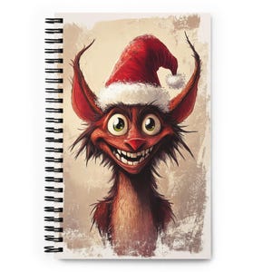 May include: Spiral-bound notebook featuring a whimsical illustration of a red creature with large ears, a Santa hat, and a wide grin. The artwork has a cartoonish style with a textured background, perfect for jotting down notes or sketches.