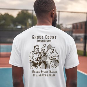 May include: White t-shirt featuring a tennis-themed graphic with the text "Ghoul Court Tennis Center." The graphic depicts Frankenstein's monster, a skeleton, and a ghost playing tennis. The text "Where Every Match Is A Grave Affair" is also included.