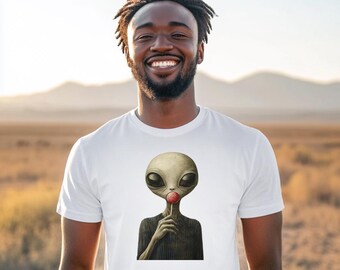 May include: White t-shirt featuring a graphic of a gray alien holding a red lollipop. The alien has large black eyes and is depicted in a dark gray sweater. The background is a blurred outdoor scene.