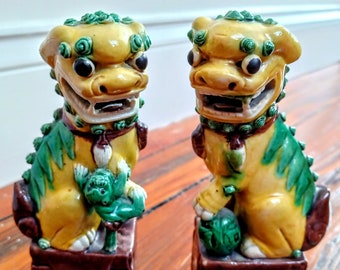May include: A pair of yellow and green ceramic foo dogs, traditional Chinese guardian lions, with intricate details and a glazed finish. Each foo dog is standing on a brown base.