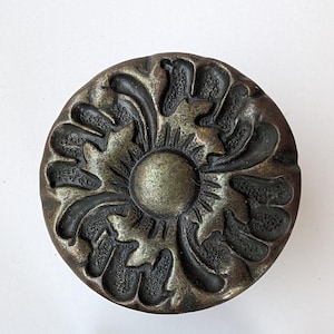 May include: A circular metal cabinet knob with a dark bronze finish. The surface features a raised, radiating floral pattern with a smooth central dome and textured, petal-like segments extending toward the outer edge of the round knob.