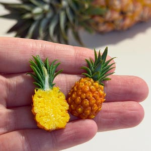 Pode incluir: Duas esculturas de abacaxi em miniatura, uma cortada ao meio para mostrar o interior amarelo, a outra inteira. Ambas têm copas verdes e frondosas e são seguradas em uma mão. Um abacaxi maior está ao fundo.