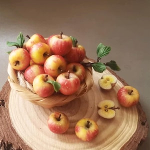 May include: A collection of small, red and yellow apples with green leaves, some whole and some sliced, are displayed in a woven basket and on a wooden slice. The apples have a realistic appearance, suggesting they are decorative or for display.