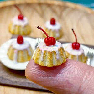 Puede incluir: Mini pastelitos dorados con glaseado blanco, chispas y una cereza roja brillante encima. Varios pasteles están dispuestos en un plato blanco pequeño y una superficie de madera. Los pasteles son sostenidos por un dedo.