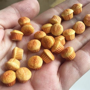 Puede incluir: Una colección de cupcakes en miniatura con tapas doradas y envoltorios de papel naranja. Los cupcakes están dispuestos en una mano, mostrando su pequeño tamaño y diseño detallado. La imagen destaca la apariencia realista de los productos horneados.