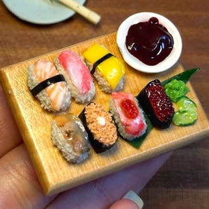 以下が含まれることがあります： 木製のサービングボードにミニチュア寿司セット。寿司には、サーモン、マグロ、その他のカラフルなトッピングが含まれています。小さな白いボウルには、濃い色のディップソースが入っています。緑色のワサビとキュウリのスライスがプレゼンテーションを完成させます。