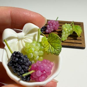 Puede incluir: Un cuenco de cerámica blanca en miniatura con forma de lazo que contiene tres pequeños racimos de uvas artificiales en tonos morado oscuro, verde claro y rosa, cada uno con una hoja verde, junto a dos racimos de uvas en una bandeja marrón.
