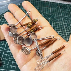Puede incluir: Una colección de utensilios de cocina en miniatura que incluye batidores de alambre, cucharas ranuradas y coladores de malla. Cada herramienta tiene una cabeza de metal plateado y un mango de madera marrón, con cuentas de madera verde oliva.