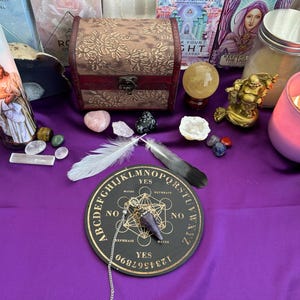 May include: A black and gold Ouija board with a purple pendulum and a white and black feather on a purple cloth. The board has the letters of the alphabet, numbers, and the words "yes", "no", and "goodbye".