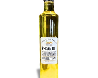 May include: A clear glass bottle of 100% pure pecan oil from The Pecan Press. The label is white with black text and features a pecan nut design. The bottle is 500ml (16 fl oz) and is made in Powell, Texas.