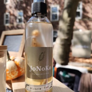 May include: A clear glass spray bottle with a black and clear plastic top. The bottle has a tan label with the words "JoNoKo" and "Muscle Tea". The liquid inside is a light yellow color. The background includes a tree and a brick building.
