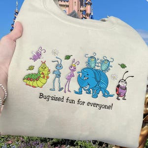 May include: Cream-colored sweatshirt featuring a colorful embroidered design of cartoon insects. The design includes a caterpillar, ants, a rhino-like bug, and a ladybug. The text "Bug-sized fun for everyone!" is below the design.