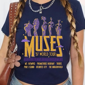 May include: Navy blue t-shirt featuring a graphic of the Muses from Greek mythology. The design includes the word "MUSES" in gold, the text "'97 WORLD TOUR", and various locations. The shirt has a classic crew neck.