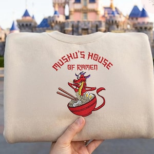 Op de afbeelding: Beige sweatshirt met de rode tekst "Mushu's House of Ramen". Daaronder een geborduurd beeld van Mushu de draak die stokjes boven een kom ramen vasthoudt. De achtergrond is wazig.