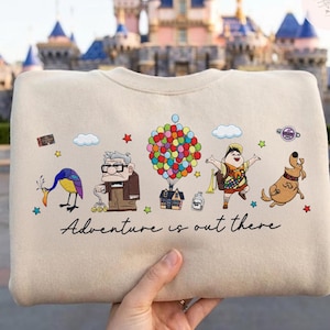 May include: Beige crewneck sweatshirt featuring a colorful graphic of characters and elements from the animated film "Up." The design includes the text "Adventure is out there." The background shows a castle.
