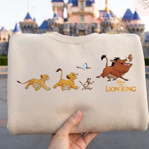 May include: A cream-coloured sweatshirt with an embroidered design from Disney's The Lion King. The design features Simba, Timon, Pumbaa, and a bird, with the words "The Lion King" in gold lettering. The sweatshirt is held by a person.