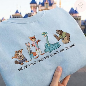 May include: Light blue sweatshirt featuring embroidered cartoon characters from a popular animated film. The design includes a beaver, rabbit, fox, snake, and sloth. The text "WE'RE WILD AND WE CAN'T BE TAMED" is also embroidered on the sweatshirt.