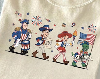 Puede incluir: Camiseta color crema con un desfile patriótico de personajes de dibujos animados. El diseño incluye un baterista, un vaquero, una vaquera y un dinosaurio sosteniendo una bandera. Fuegos artificiales y estrellas decoran la parte superior del diseño. La camiseta está doblada sobre unos vaqueros.