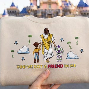 May include: Beige sweatshirt with a graphic of a figure in a white robe and yellow shawl, holding hands with a cartoon cowboy and Buzz Lightyear. The text "YOU'VE GOT A FRIEND IN ME" is printed below the figures. Stars, clouds, and parachuters are also featured.