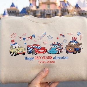 Pode incluir: Camisola bege com carros de desenho animado com designs patri&oacute;ticos e fogos de artif&iacute;cio. O texto "Happy 250 Years of freedom 1776-2026" est&aacute; impresso abaixo dos carros. A camisola &eacute; segurada por uma pessoa.