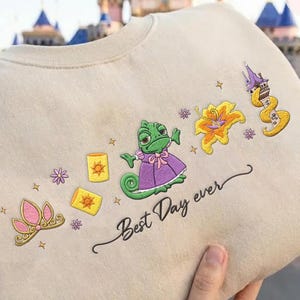 May include: Beige sweatshirt with embroidered cartoon graphics of a green chameleon, a pink tiara, yellow lanterns, and a yellow flower. The text "Best Day ever" is written in black script.